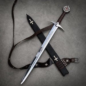 Puede incluir: Una espada de metal plateado con empuñadura forrada en cuero marrón y un pomo circular con un emblema de cruz roja. La espada descansa diagonalmente sobre una vaina de cuero negro y un cinturón largo de cuero con hebilla metálica.