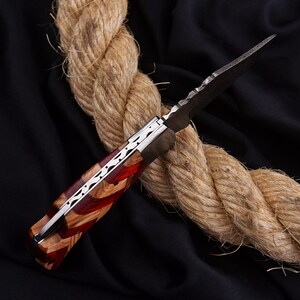 Puede incluir: Un cuchillo artesanal con una hoja oscura y un mango con un dise&ntilde;o de grano de madera y rojo. El cuchillo tiene un acento plateado y est&aacute; sobre una cuerda gruesa sobre un fondo negro.