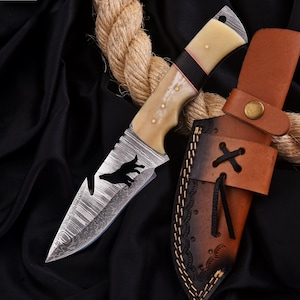 Puede incluir: Un cuchillo de acero de Damasco con mango de hueso, detalles negros y una funda de cuero. La hoja presenta la silueta de un lobo. La funda es de cuero marrón con costuras decorativas y cierre de botón. Una cuerda está en el fondo.