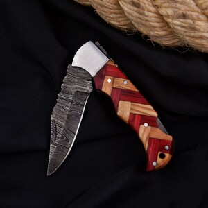 Puede incluir: Una navaja plegable con una hoja estampada y un mango de madera roja, marr&oacute;n claro y marr&oacute;n oscuro. La hoja tiene un dise&ntilde;o texturizado &uacute;nico. El mango est&aacute; hecho de m&uacute;ltiples piezas de madera. La navaja est&aacute; sobre un fondo negro.