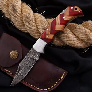 Puede incluir: Una navaja plegable con una hoja estampada y un mango de madera roja y clara. El cuchillo descansa sobre una funda de cuero marr&oacute;n con un bot&oacute;n decorativo. Una gruesa cuerda est&aacute; en el fondo.