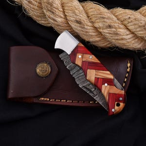 Puede incluir: Una navaja plegable con un mango de madera estampada en tonos rojos y beige, y una hoja plateada. El cuchillo descansa sobre una funda de cuero marr&oacute;n con un bot&oacute;n met&aacute;lico decorativo. Una cuerda gruesa est&aacute; en el fondo.