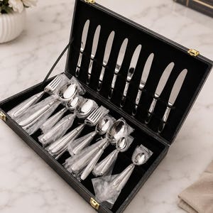 May include: A black rectangular storage case with gold-tone latches containing a set of polished stainless steel flatware. The set includes knives arranged in the lid and various spoons and forks wrapped in clear plastic within the base.