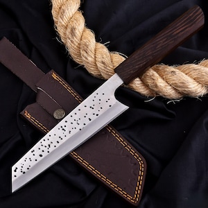 Puede incluir: Un cuchillo de chef de acero inoxidable con mango de madera oscura y funda de cuero. La hoja tiene una serie de pequeñas perforaciones espaciadas. La funda marrón tiene costuras amarillas y un cierre de botón metálico.