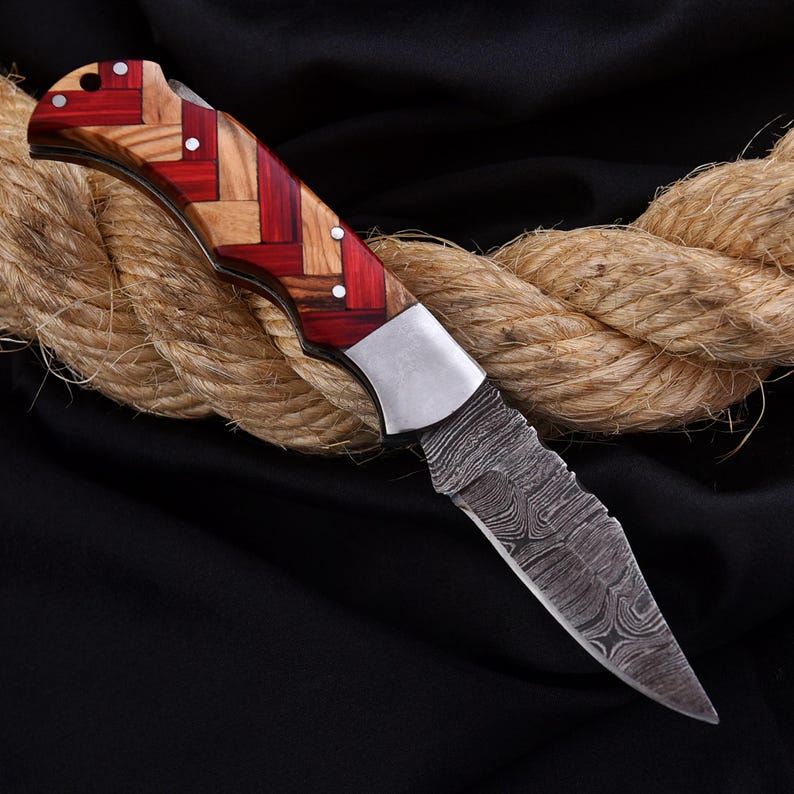 Puede incluir: Una navaja plegable con una hoja estampada y un mango de madera roja y clara. La hoja tiene un dise&ntilde;o texturizado &uacute;nico. El cuchillo est&aacute; apoyado sobre una gruesa cuerda sobre un fondo negro.