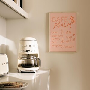 Puede incluir: Cafetera SMEG color crema con una jarra de vidrio llena de caf&eacute;. Un letrero rosa enmarcado muestra "CAFE PSALM" y un men&uacute; de bebidas y comida. Tambi&eacute;n se ven una tostadora SMEG y un plato de tostadas.