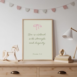 Puede incluir: Un cuadro rectangular con marco que presenta un fondo beige claro, un lazo rosa dibujado con líneas en la parte superior y el texto en verde oscuro "She is clothed with strength and dignity. Proverbs 31:25" centrado en una fuente serif simple.
