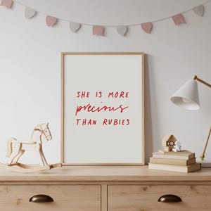 May include: A rectangular wall art print in a light wood frame featuring the red text "SHE IS MORE precious THAN RUBIES" centered on a white background. The print is displayed on a wooden surface alongside a wooden rocking horse and books.