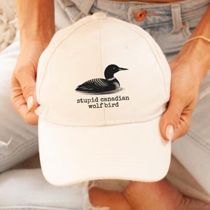 May include: A cream-colored baseball cap featuring a black and white illustration of a loon on water. The text "stupid canadian wolfbird" is printed below the bird. The cap has a curved brim and a classic six-panel design.