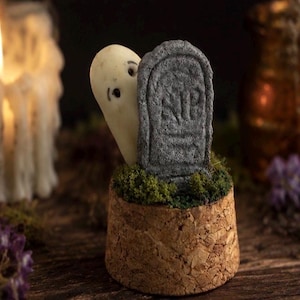 May include: A small decorative piece featuring a white ghost figurine peeking from behind a grey tombstone with the letters RIP engraved on the front. The figures are set into a cylindrical cork base topped with a layer of green moss.