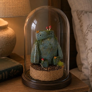Peut inclure: Une figurine bleu-vert rappelant une pierre, ornée de mousse et de petits champignons rouges, repose sur une base en liège circulaire sous une cloche en verre transparent avec un rebord en bois sombre. Elle a une forme cubique et deux yeux.