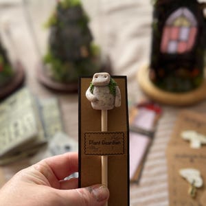 May include: A small, speckled, off-white clay figure with green moss-like accents attached to a wooden stick. The figure is mounted on a brown rectangular card featuring the text Plant Guardian, which is displayed on a dark brown backing.