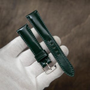 May include: A dark green leather watch strap featuring a smooth, glossy finish with tonal stitching along the edges. The strap includes a silver-toned metal buckle and a matching leather keeper loop, designed for secure attachment to a timepiece.