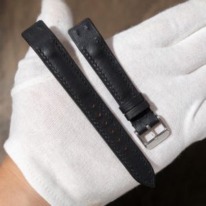 May include: Black leather watch strap with a silver buckle. The strap has detailed stitching along the edges and several holes for adjustment. The watch band is shown against a white background.