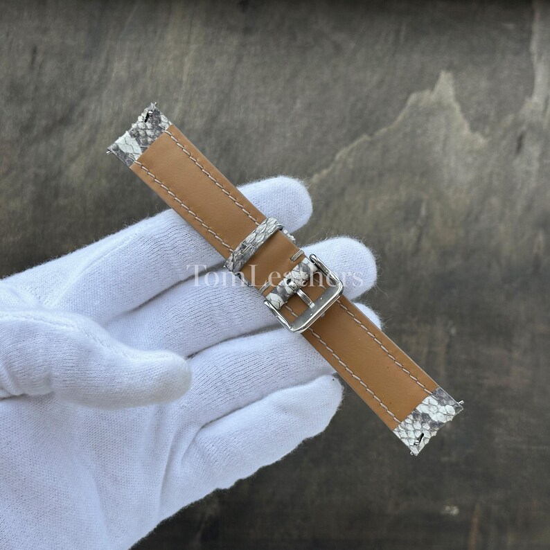 May include: A watch strap featuring a tan leather center with white stitching and grey and white snakeskin patterned leather ends. The strap includes a silver-toned metal buckle and a matching snakeskin patterned leather keeper loop.