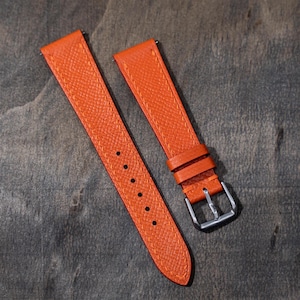 以下が含まれることがあります： オレンジ色の2ピースの時計ストラップ。表面はテクスチャ加工され、オレンジ色のステッチが施されています。ストラップにはシルバー色のバックルといくつかの調整穴があります。時計バンドは革製です。