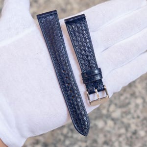 Peut inclure: Deux bracelets de montre bleu marine avec un motif texturé en forme de reptile. Un bracelet a une boucle dorée. Les bracelets sont présentés sur une surface blanche. Les bracelets de montre sont une pièce de rechange pour une montre.