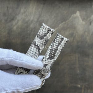 May include: A watch strap featuring a textured snakeskin pattern in shades of grey, white, and black. The strap consists of two pieces with visible stitching along the edges and a metal buckle component at the base, showcasing a detailed scale design.