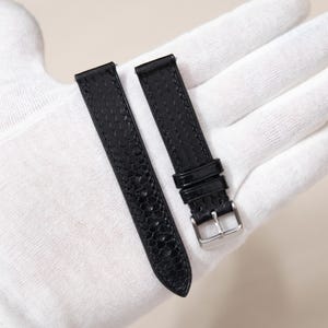 May include: Two black watch straps with a textured, snake-like pattern. One strap features a silver buckle. The straps are shown on a white surface, possibly a gloved hand. The watch straps are new.