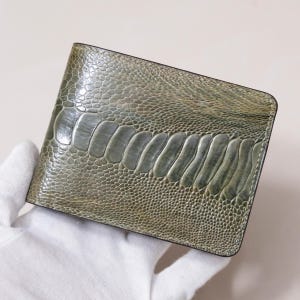 以下が含まれることがあります： オリーブグリーンの質感のあるダチョウ革の二つ折り財布。財布の中央には大きめの鱗のような模様があり、縁は滑らかで濃い色です。手袋をはめた手で財布を持っています。
