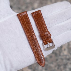 May include: Two brown watch straps with a textured, reptile-like pattern. The straps are made of leather and have a silver-coloured buckle. The watch straps are displayed on a white surface.