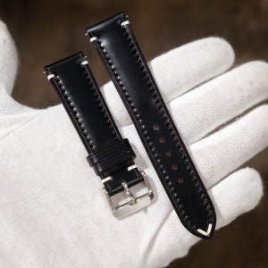 May include: Black leather watch strap with white stitching. The strap features a silver-colored buckle and is shown against a white background. The strap is designed for a watch and is made of a durable material.