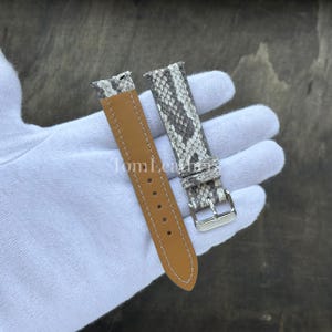 May include: A two-piece watch strap featuring a grey and white snakeskin pattern on the outer side and a smooth tan leather lining on the inner side. The strap includes a silver-toned metal buckle and white stitching along the edges.