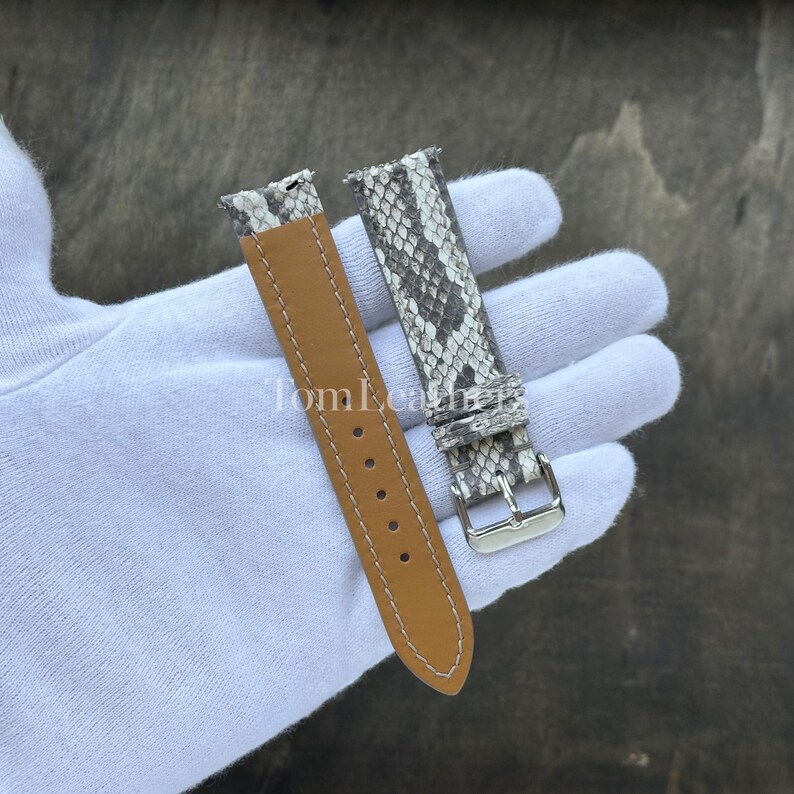 May include: A two-piece watch strap featuring a grey and white snakeskin pattern on the outer side and a smooth tan leather lining on the inner side. The strap includes a silver-toned metal buckle and white stitching along the edges.