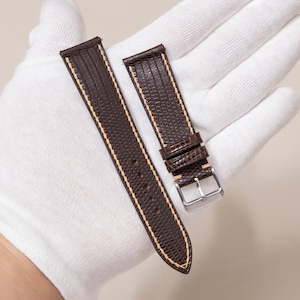 May include: A pair of brown watch straps with a textured, reptile-like pattern. The straps have cream-colored stitching along the edges and a silver-colored buckle. The watch straps are displayed on a white surface.
