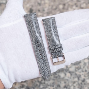 May include: Two grey leather watch straps with a textured pattern. One strap has a gold-coloured buckle. The straps are displayed on a white surface.