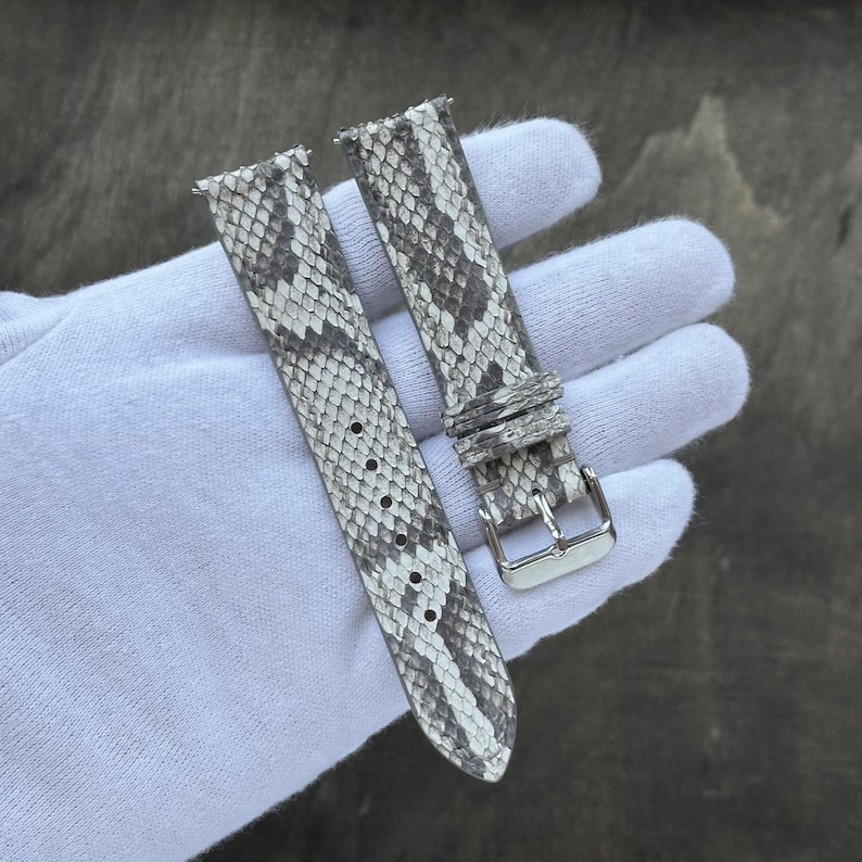 May include: A two-piece watch strap featuring a grey and white snakeskin pattern. The strap includes a silver-toned metal buckle and two matching sliding keepers. The surface texture displays a detailed, repeating scale design throughout the band.