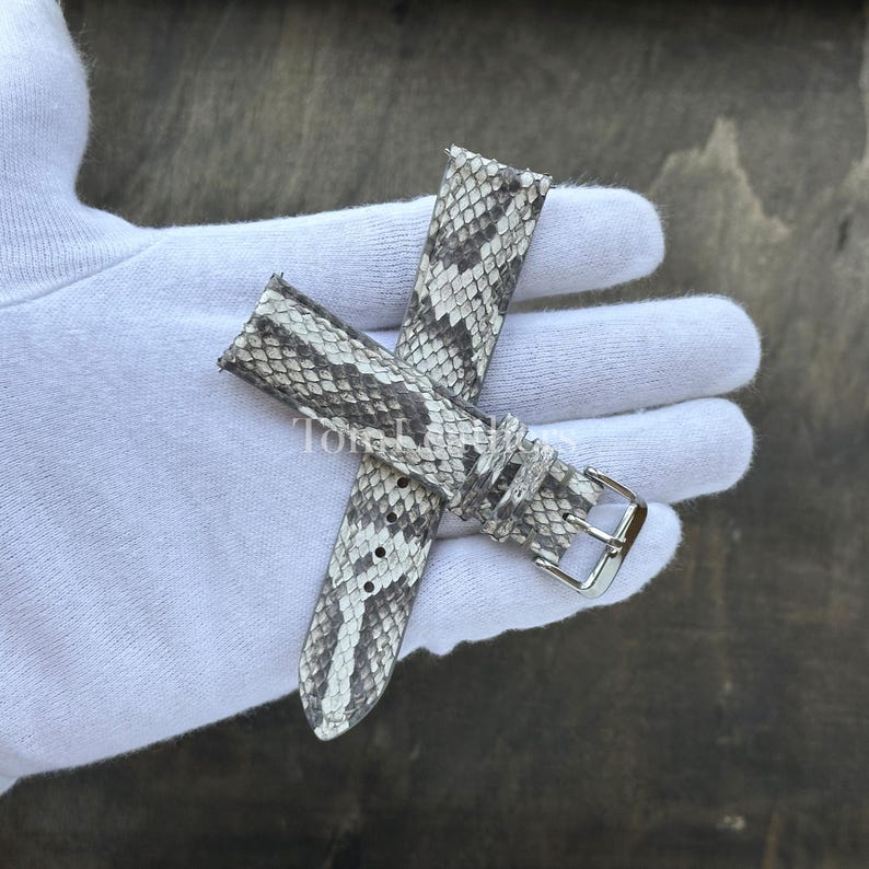 May include: A two-piece watch strap featuring a grey and white snakeskin pattern with a textured scale finish. The strap includes a polished silver-tone metal buckle and a matching sliding keeper loop, designed for secure attachment to a timepiece.