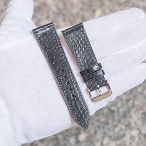 May include: A pair of dark grey watch straps with a textured pattern. The straps have a gold-coloured buckle and adjustment holes. The straps are held by a gloved hand.