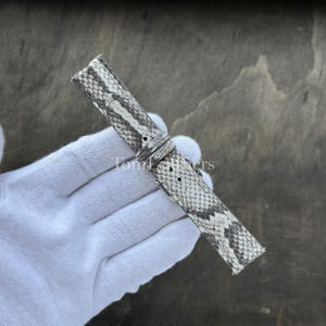 May include: A watch strap featuring a grey and white snakeskin pattern with a textured scale finish. The strap includes a matching sliding keeper loop and a series of pre-punched holes for adjustment, showcasing a detailed reptilian aesthetic.