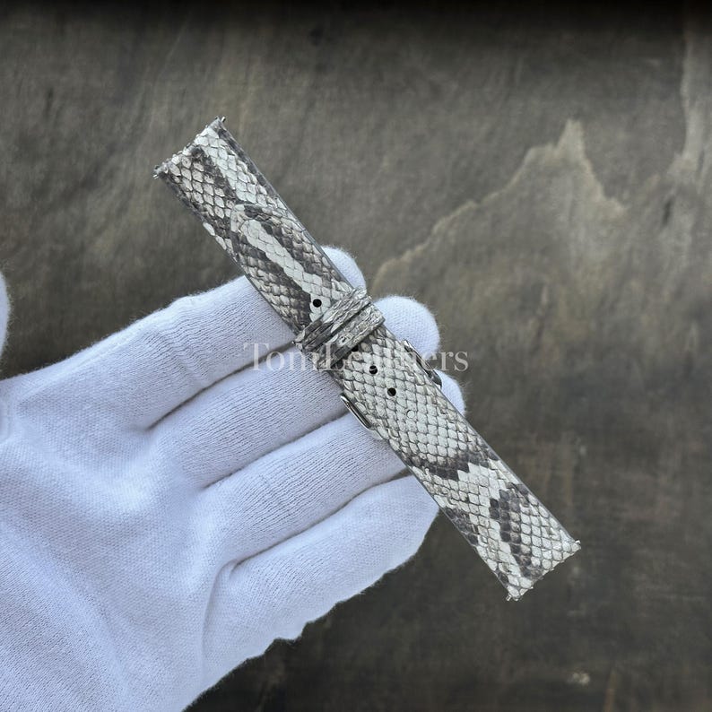 May include: A watch strap featuring a grey and white snakeskin pattern with a textured scale finish. The strap includes a matching sliding keeper loop and a series of pre-punched holes for adjustment, showcasing a detailed reptilian aesthetic.