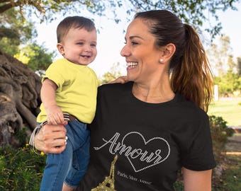 May include: A woman wearing a black t-shirt with a rhinestone design that says "Amour" with a heart and "Paris, Mon Amour" underneath. She is holding a young child in her arms. The child is wearing a yellow t-shirt and blue jeans.