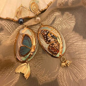 Puede incluir: Un par de pendientes ovalados con diseños de alas de mariposa en tonos azules, naranjas y negros. Los pendientes tienen detalles dorados y un dije dorado en forma de cola. También se adjuntan una perla y una cuenta azul oscuro.
