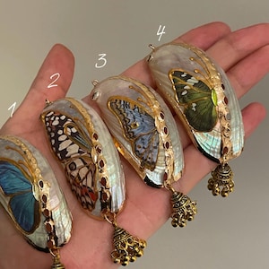 May include: Four butterfly wing pendants with unique designs. Each pendant showcases a different butterfly pattern in blue, brown, and green hues, set against a pearlescent backdrop. Gold-coloured metal detailing and a bell charm complete the design.