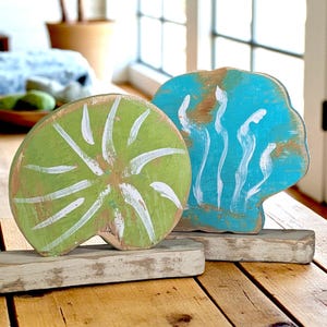 May include: Two wooden decorative pieces. One is a green, circular shape with white brushstrokes. The other is a blue, cloud-like shape with white and brown accents. Both are mounted on rectangular wooden bases. The pieces are on a wooden table.