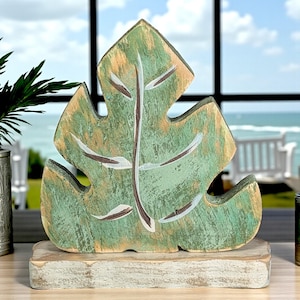 May include: A wooden leaf sculpture with a distressed green and tan finish, mounted on a rectangular base. The leaf features white and brown painted details, creating a tropical aesthetic. The sculpture is a decorative accent piece.