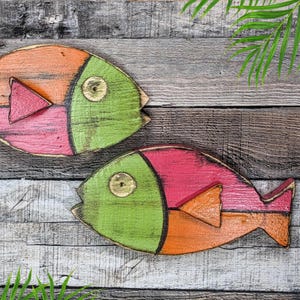 May include: Two wooden fish painted in bright colors. The fish are green, orange, and pink. They are hanging on a wooden wall.