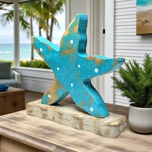 May include: A large, turquoise wooden starfish sculpture with gold accents and white polka dots. The starfish is mounted on a rectangular, weathered, off-white base. The sculpture is a decorative coastal accent.