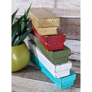 May include: A stack of six rectangular wooden boxes painted in various colors: gold, red, green, white, and turquoise. The boxes are arranged in a staggered formation, with the gold box at the top. A green plant in a yellow-green pot is on the left.
