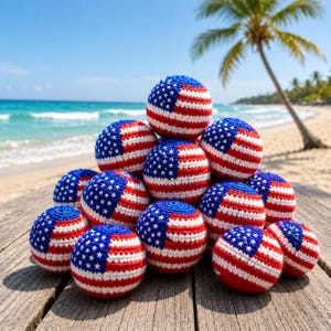 May include: A collection of crocheted balls, each meticulously crafted to resemble the American flag. The balls feature a blue field with white stars and alternating red and white stripes. They are arranged on a weathered wooden surface with a beach and ocean backdrop.