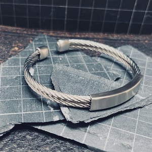 May include: A silver cuff bracelet with a braided cable design. The bracelet has a rectangular bar in the center and rounded caps at the ends. It is displayed on a dark grey surface. The bracelet's design is modern.