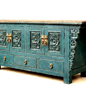 May include: A rectangular wooden sideboard cabinet finished in a distressed teal blue paint. The piece features four cabinet doors with carved floral panels and brass hardware, plus three lower drawers with circular brass pulls and ornate side brackets.