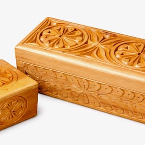 Könnte beinhalten: Zwei Holzkisten mit aufwendigen Blumenschnitzereien. Die größere Kiste ist rechteckig, die kleinere quadratisch. Beide sind aus hellem Holz gefertigt und weisen detaillierte, symmetrische Designs auf Deckeln und Seiten auf.