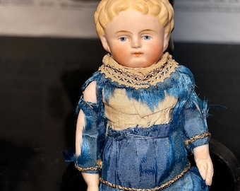 May include: A vintage porcelain doll with blonde hair and blue eyes. The doll wears a blue dress with gold trim and a tan bodice. The doll's dress has a distressed, aged appearance, suggesting it is antique.