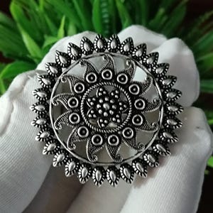 May include: A silver-colored ring with an intricate, circular design. The ring features a central floral pattern surrounded by a series of smaller circles and teardrop-shaped embellishments. The ring is held against a white background.