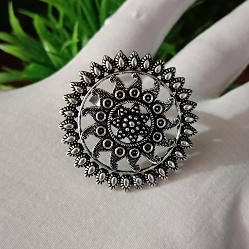 May include: A silver-colored statement ring with a detailed, sunburst design. The ring features a central floral pattern surrounded by circular and teardrop-shaped elements. The ring is displayed on a white surface.